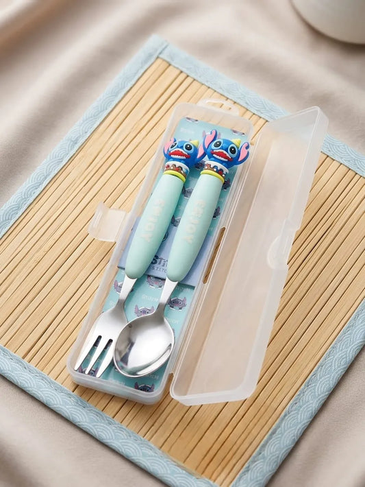 A children's stainless steel spoon and fork set in a clear plastic case, featuring mint green handles with cartoon character decorations, placed on a bamboo mat.