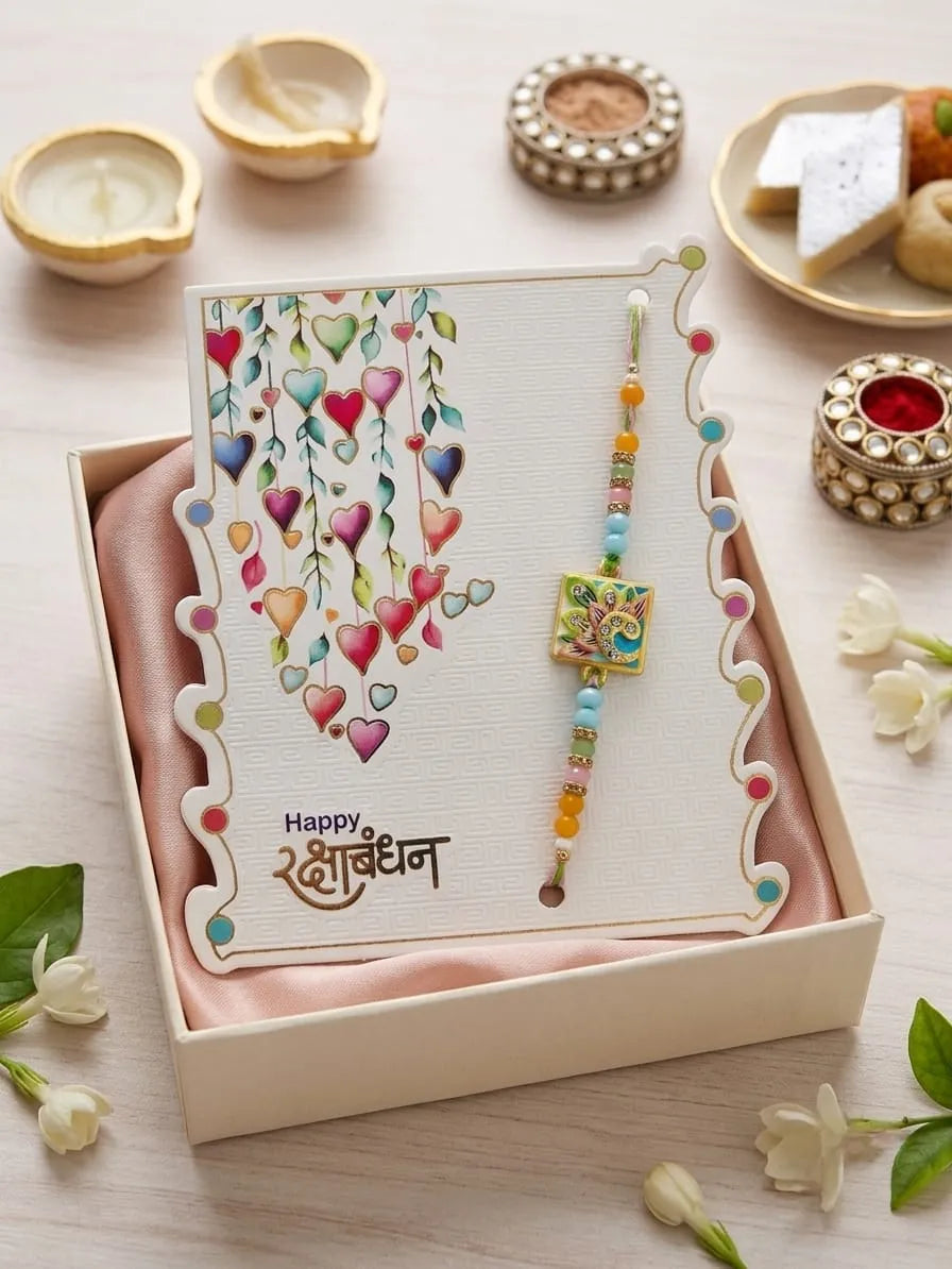 A boxed Rakhi gift set featuring a decorative card with colorful hanging hearts and the text 'Happy Rakhi' in Devanagari script, alongside a beaded Rakhi with a square floral charm. The set is presented in an open cream-colored box lined with pink fabric, surrounded by festive props on a light wooden surface.