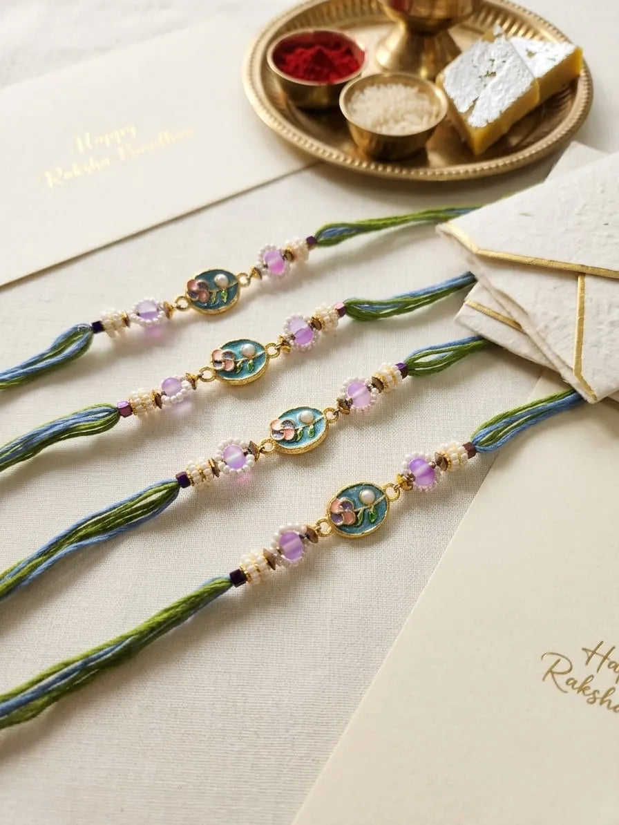 Four traditional Indian Rakhi bracelets laid out on a cream fabric surface, decorated with green and blue threads, gold-toned beads, and pink stones. Cards reading 'Happy Raksha Day' and a decorative gold tray with sweets are visible in the background.