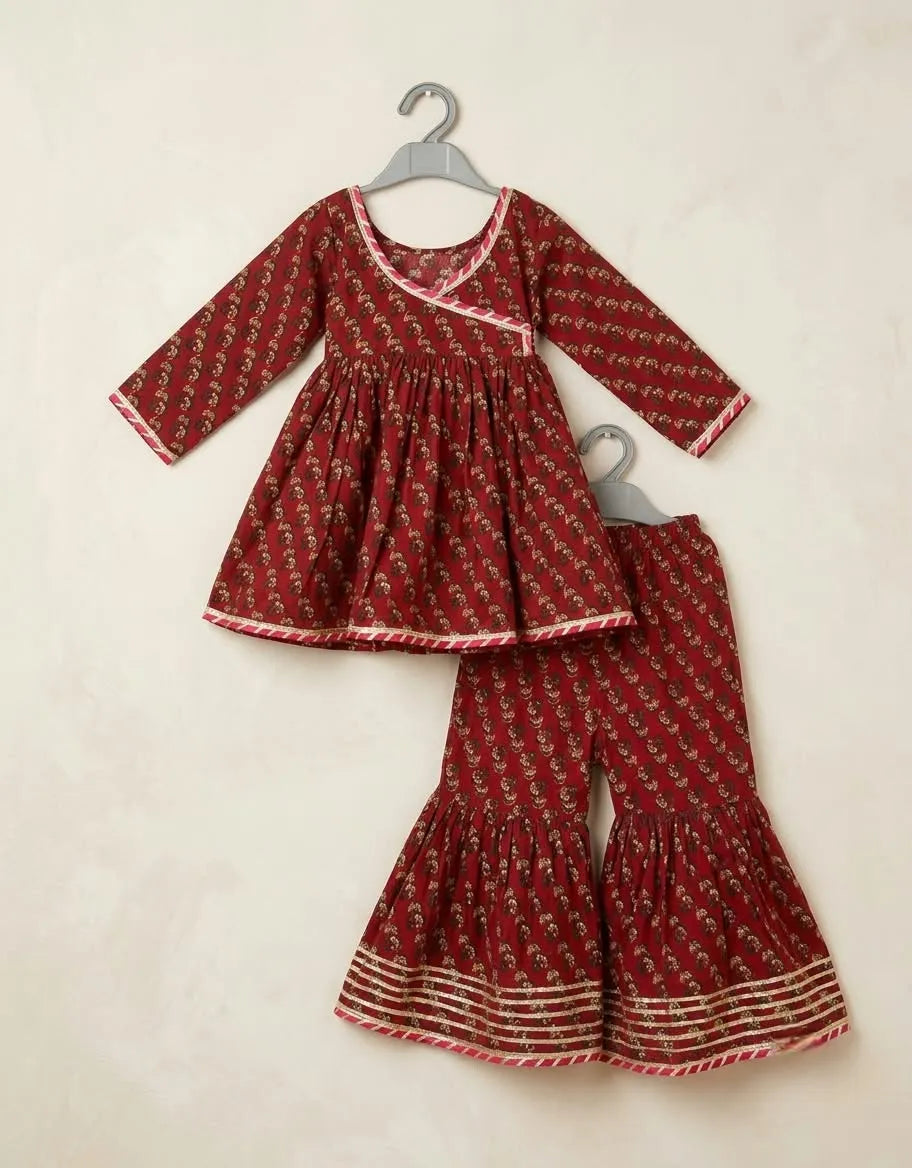 A two-piece toddler girl's outfit set consisting of a long-sleeve wrap-style top and matching flared pants, both in a deep red fabric with a small white floral print and white border trim, hanging on gray hangers against a light beige textured wall.