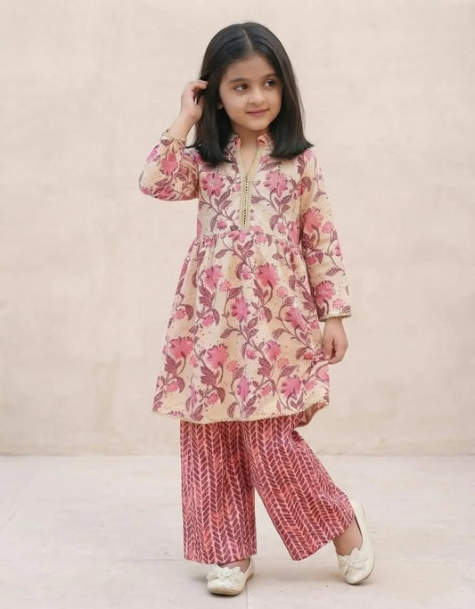 A young girl modeling a two-piece traditional outfit consisting of a beige tunic with a pink floral print and matching patterned wide-leg pants, set against a plain beige wall.