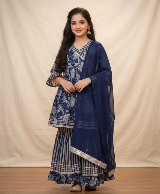 A woman modeling a traditional Indian outfit consisting of a navy blue printed kurta, matching striped sharara pants, and a sheer navy dupatta, set against a neutral draped backdrop.