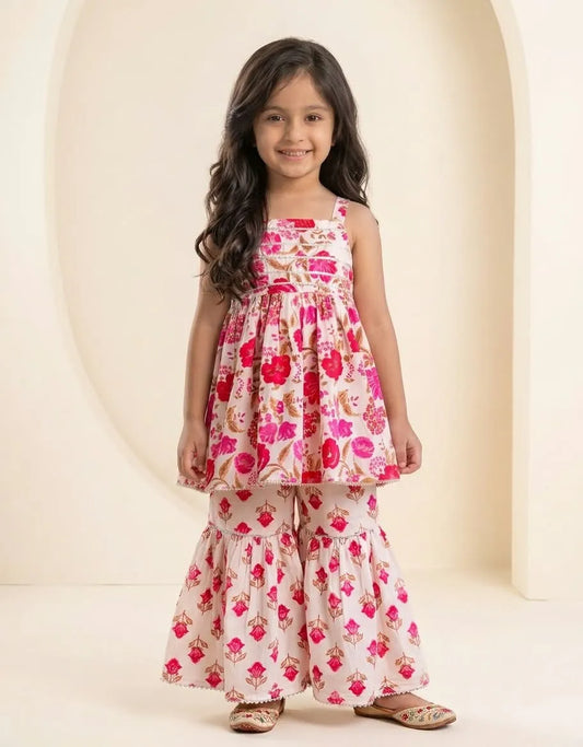 A young girl with long dark hair stands in a studio wearing a two-piece floral print outfit consisting of a sleeveless tiered top and matching wide-leg pants. The outfit has a pink, red, and green floral pattern on a cream background.
