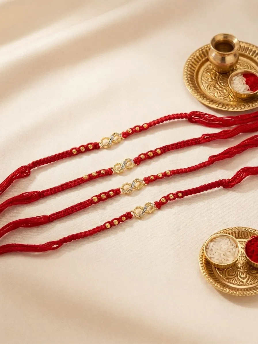 Four red braided Rakhi cords with gold and white decorative beads, arranged on a cream satin fabric alongside traditional gold-toned diya props.