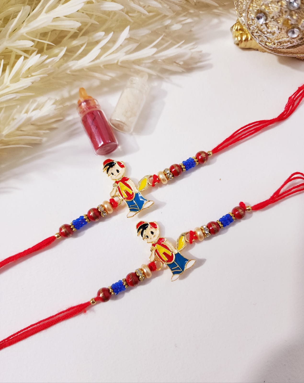 Two kids' rakhis with red and blue beads, featuring small figures of cartoon characters, presented alongside a small container and decorative feather.