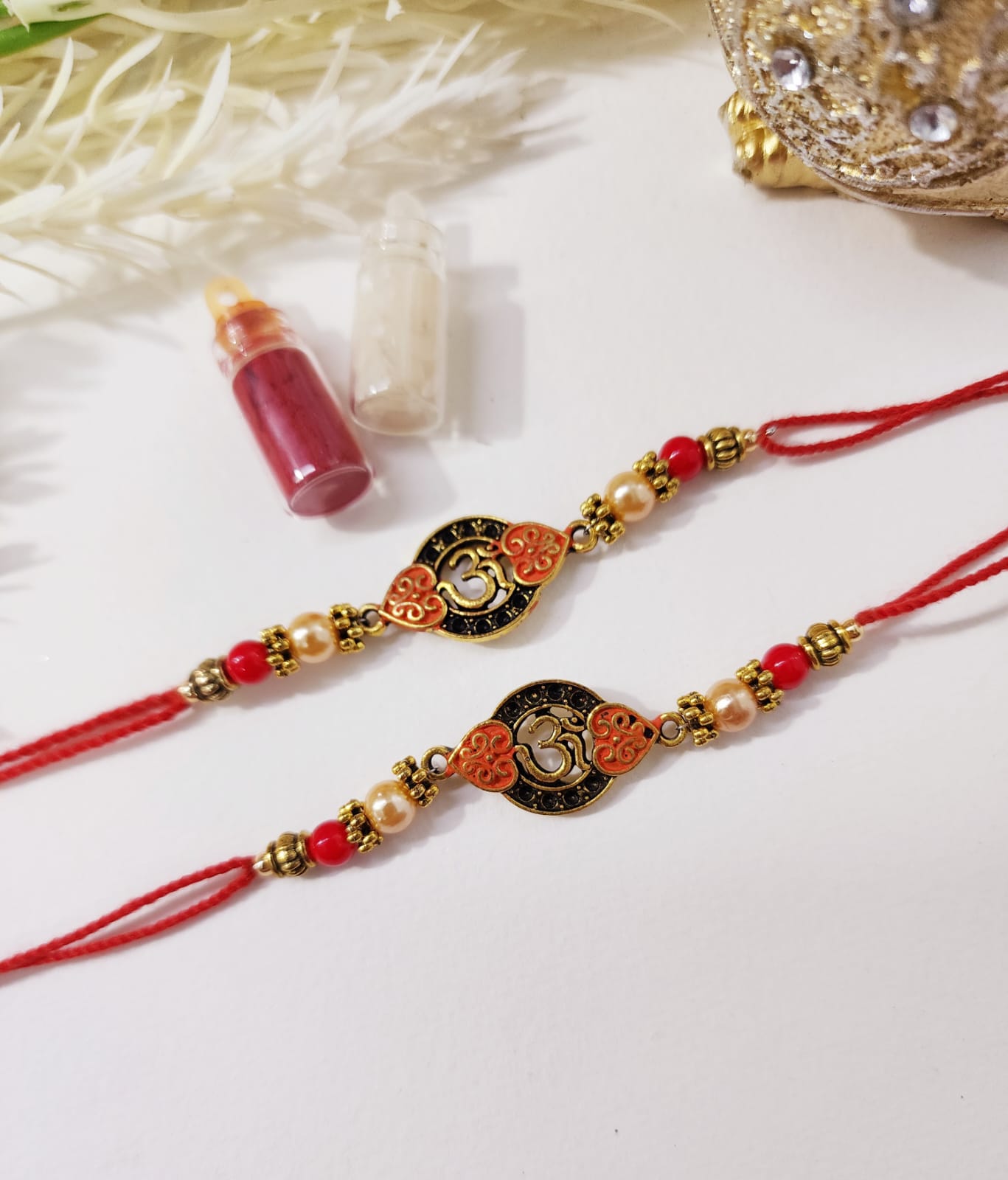 Two traditional Indian rakhi bracelets with red thread and decorative elements, displayed alongside a small bottle of tilak (a ceremonial mark).