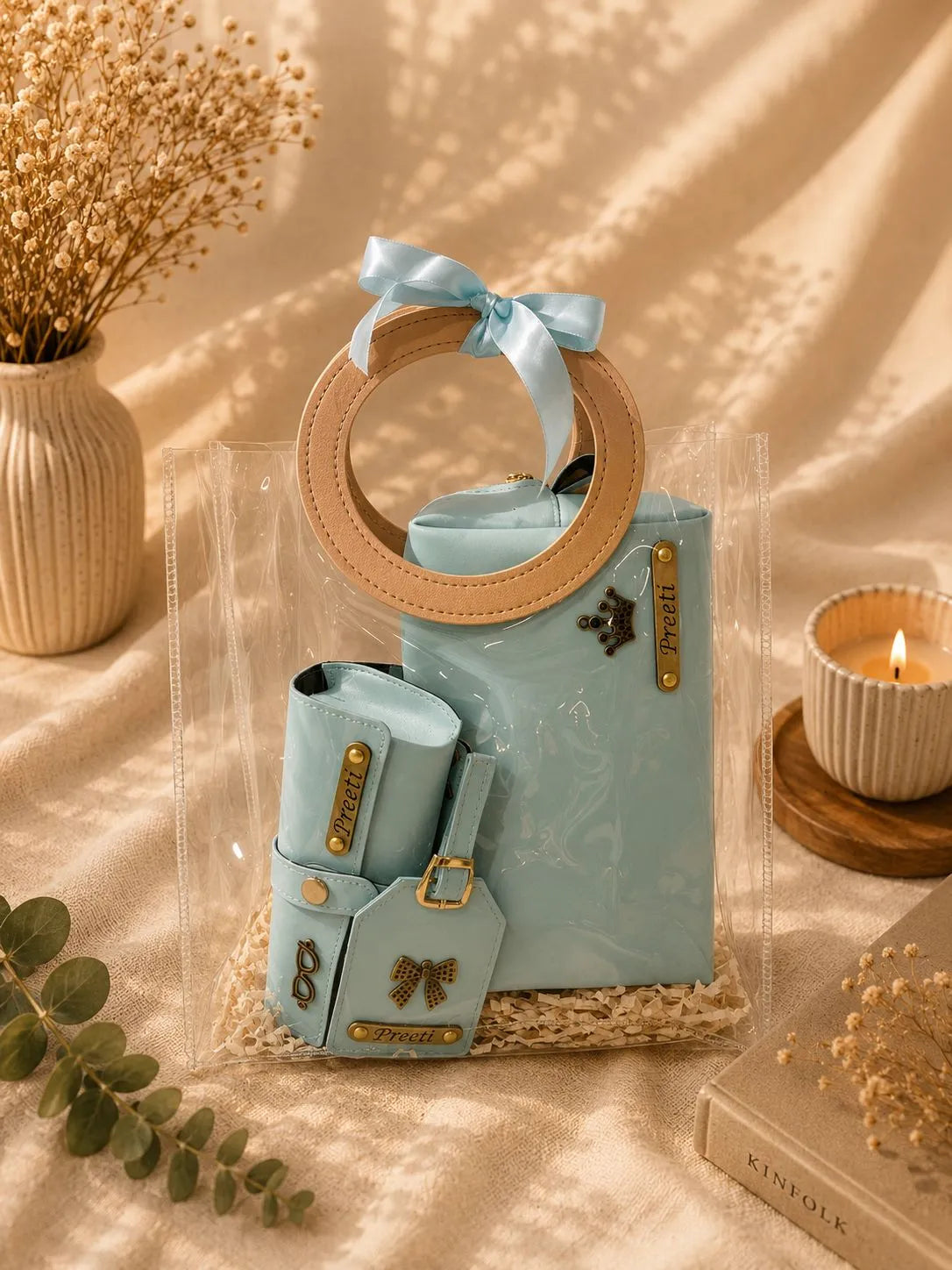 A light blue faux-leather handbag set styled in a warm, bohemian setting. The set includes a main handbag with a circular tan handle and a matching small pouch, both decorated with gold-tone hardware and a light blue ribbon bow, set against a draped beige fabric backdrop with dried flowers and a candle.