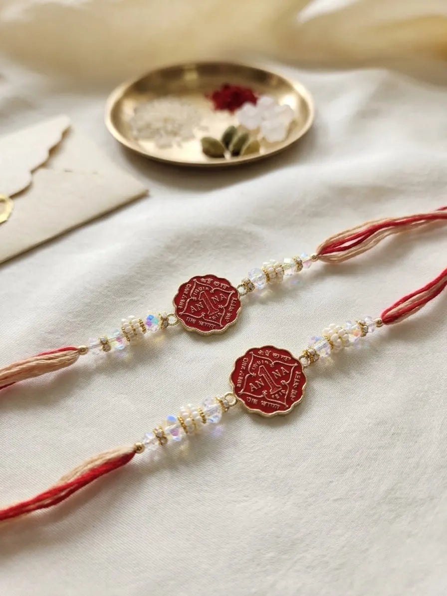 Two traditional Indian Rakhi bracelets with red cords, red circular medallions, and clear beads, displayed on a soft white fabric background with a gold dish in the background.