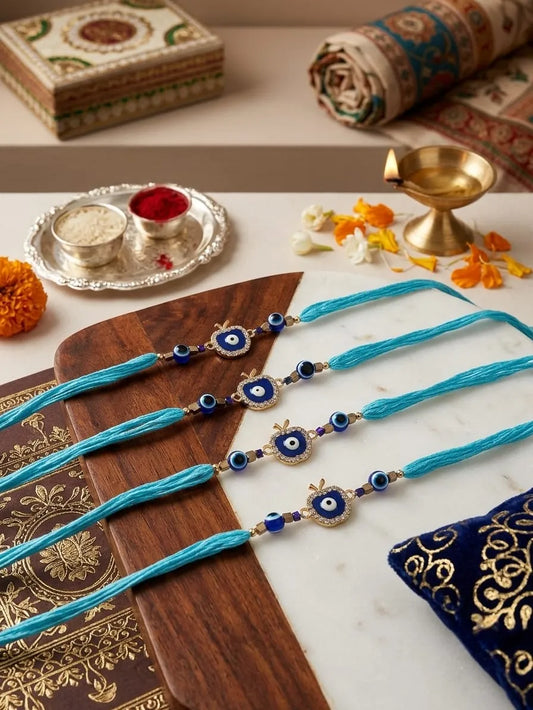 Four turquoise blue thread rakhi bracelets with blue and white evil eye charms and gold-toned borders, arranged on a wooden board over a white marble surface with festive decorative props including a brass diya, marigold petals, and patterned textiles.