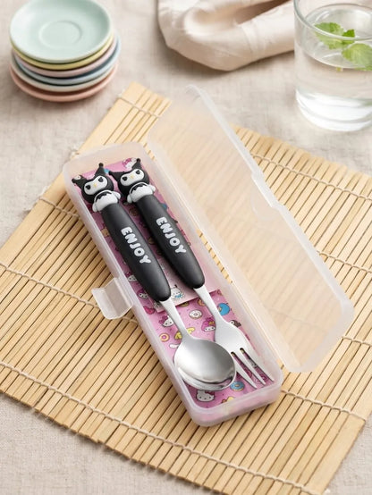 A small pink plastic utensil case holding a black-handled spoon and fork with cartoon owl faces and the word "ENJOY" on the handles, placed on a bamboo mat with stacked pastel plates and a glass of water in the background.
