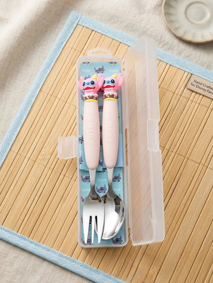 A children's stainless steel cutlery set in a clear plastic case, featuring light pink handles with cartoon character toppers and a blue patterned interior, displayed on a bamboo mat.