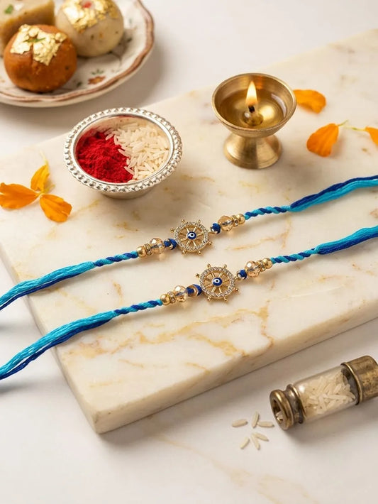 Two blue and gold braided Rakhi strands with central star-shaped evil eye beads, arranged on a white marble slab with festive props including a lit diya, red and white rice, and sweets.