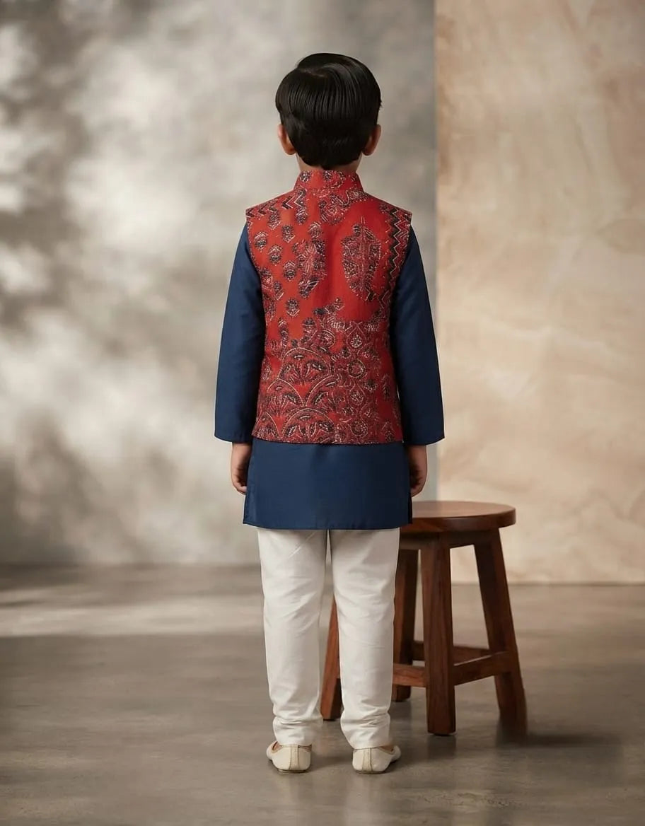 A child model shown from the back wearing a traditional Indian outfit consisting of a navy blue kurta, a red patterned Nehru jacket, and white trousers, standing next to a wooden stool in a studio setting.