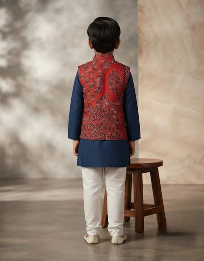 A child model shown from the back wearing a traditional Indian outfit consisting of a navy blue kurta, a red patterned Nehru jacket, and white trousers, standing next to a wooden stool in a studio setting.