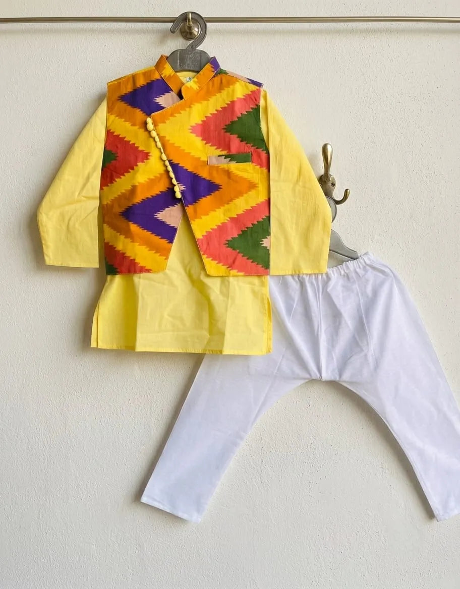 A children's traditional outfit set hanging on a wall, featuring a bright yellow kurta with a colorful geometric patterned vest, paired with white pants.