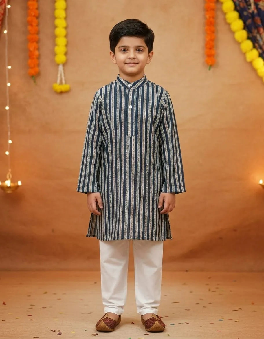 A young boy modeling a navy blue and white vertically striped kurta with white trousers and brown shoes, standing against a warm brown backdrop decorated with marigold garlands.