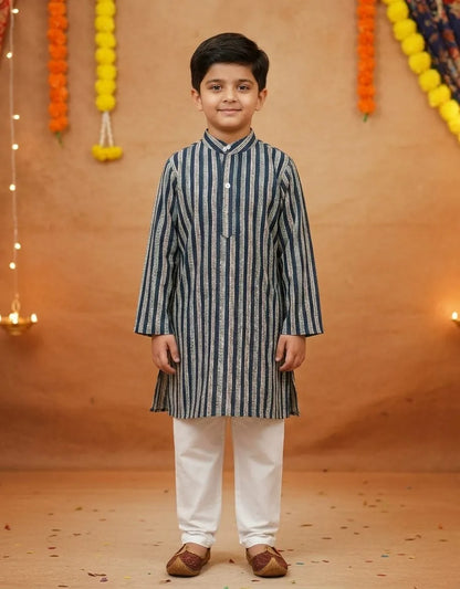 A young boy modeling a navy blue and white vertically striped kurta with white trousers and brown shoes, standing against a warm brown backdrop decorated with marigold garlands.
