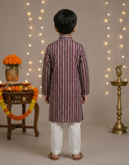 A young boy viewed from behind wearing a maroon and white vertically striped kurta with white trousers, standing in a decorated indoor setting with string lights, marigold garlands, and a brass lamp.