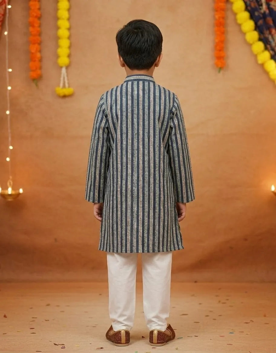 A young boy viewed from behind wearing a navy blue and white vertically striped kurta with white trousers, standing against a warm brown textured backdrop decorated with marigold garlands and string lights.