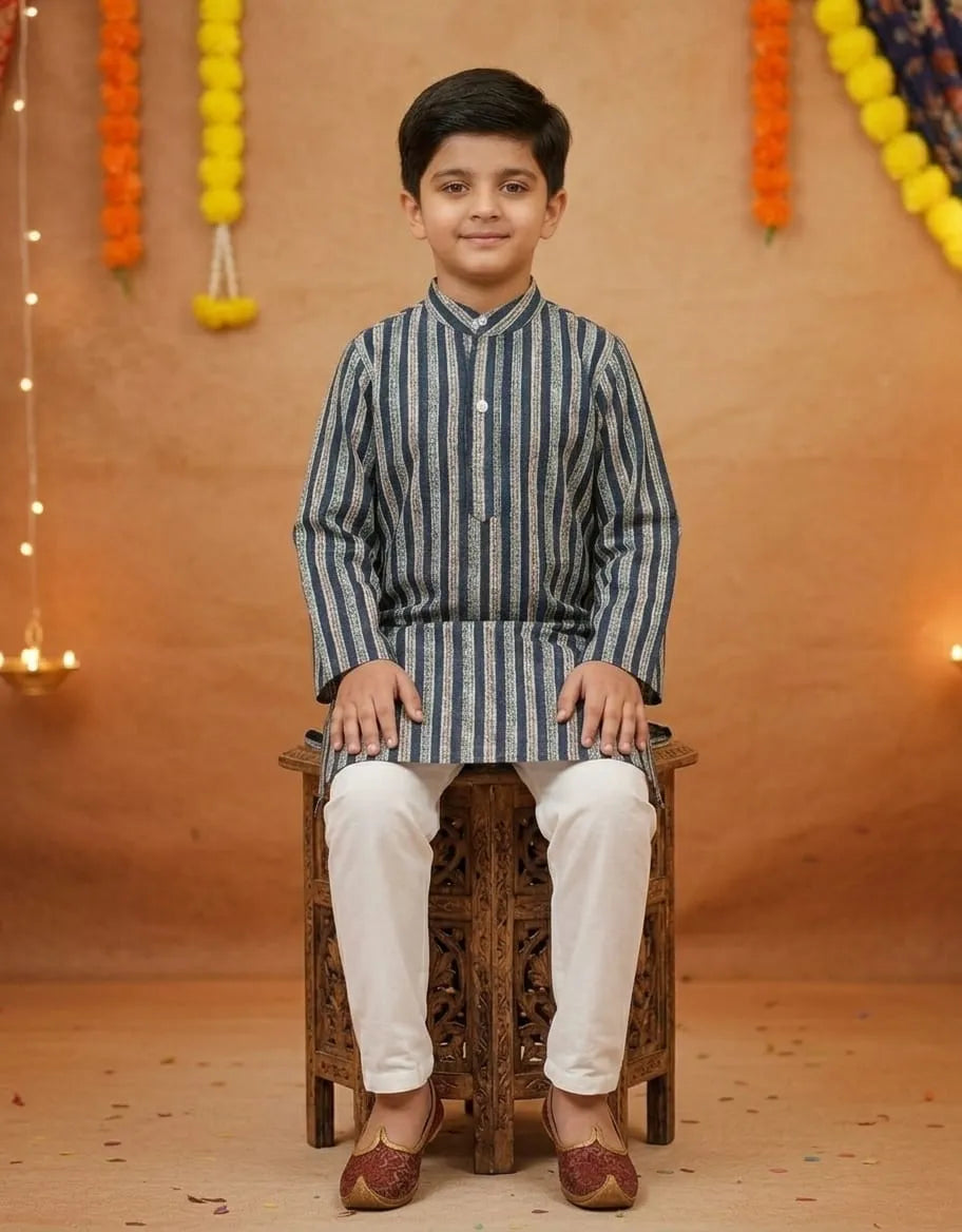 A young boy wearing a blue and white vertically striped kurta with white trousers and maroon shoes, sitting on a carved wooden stool against a warm decorated backdrop.