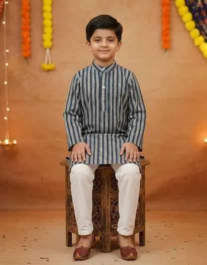 A young boy wearing a blue and white vertically striped kurta with white trousers and maroon shoes, sitting on a carved wooden stool against a warm decorated backdrop.