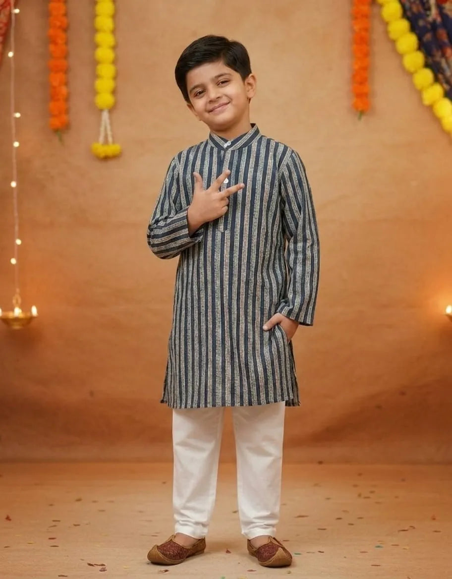 A young boy modeling a traditional Indian outfit consisting of a navy blue and white vertically striped kurta with white pants, standing against a warm brown backdrop decorated with marigold garlands.