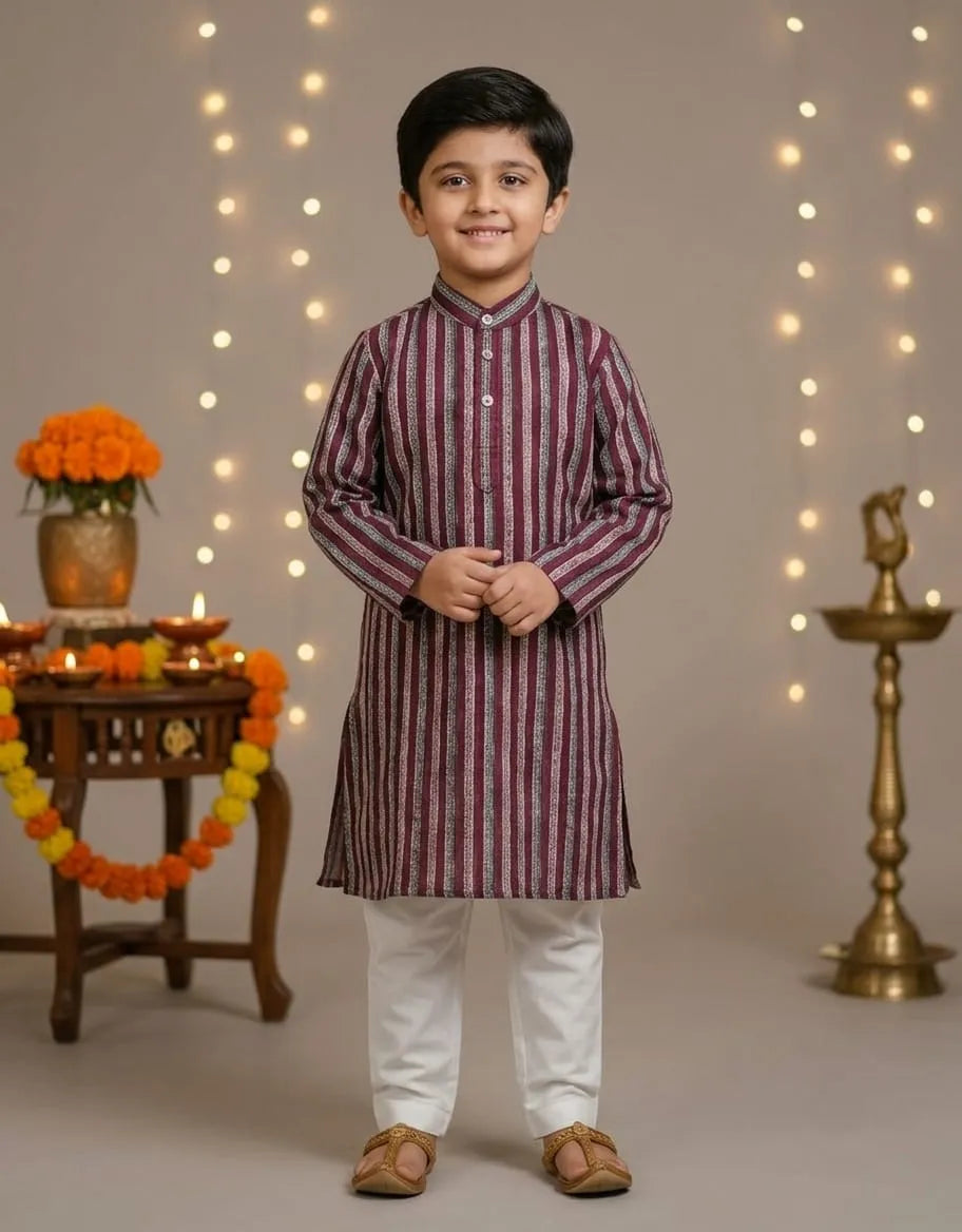 A young boy modeling a traditional Indian outfit consisting of a maroon and white vertically striped kurta with white pants, standing in front of a warm studio backdrop decorated with string lights, marigold garlands, and a brass lamp.