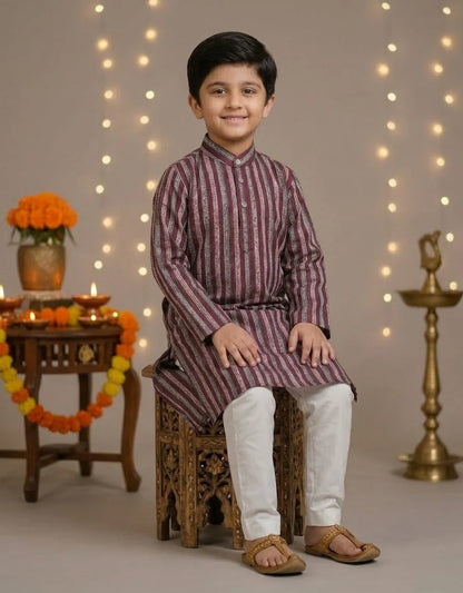 A young boy wearing a traditional Indian outfit with a maroon and white striped kurta and white trousers, sitting on a carved wooden stool in a decorated studio setting with string lights and festive props.