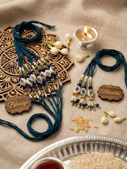 Two traditional Indian Rakhi beaded wristlets on a beige fabric background with a lit diya, white flowers, and wooden nameplates reading 'Bhaiya' and 'Bhabi'.