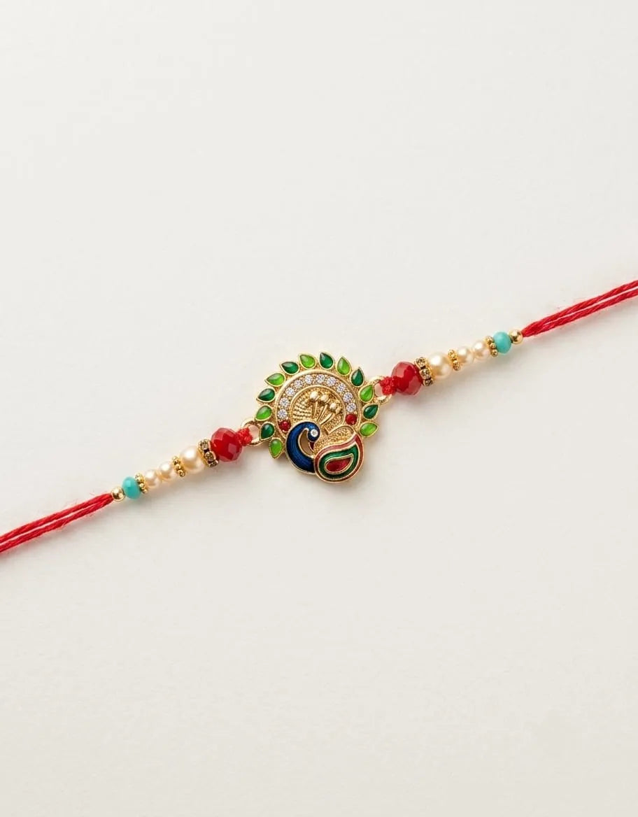 A red string rakhi bracelet featuring a central peacock charm with gold-plated details, green and blue enamel accents, and small red and turquoise beads on either side, displayed on a clean white background.