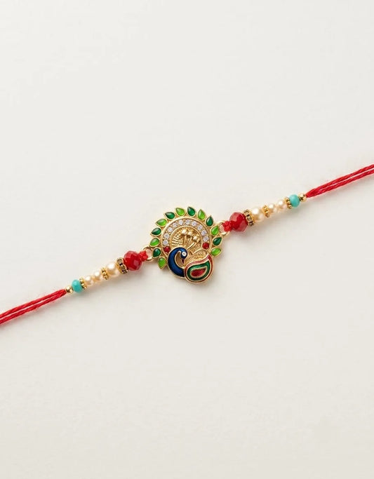 A red string rakhi bracelet featuring a central peacock charm with gold-plated details, green and blue enamel accents, and small red and turquoise beads on either side, displayed on a clean white background.