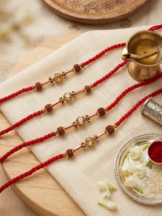 Four red braided Rakhi cords laid out on a beige cloth, each decorated with gold-toned beads and brown Rudraksha-like beads, styled with a brass pot, silver container, plate of white flowers, and a small bowl of red powder on a wooden board.