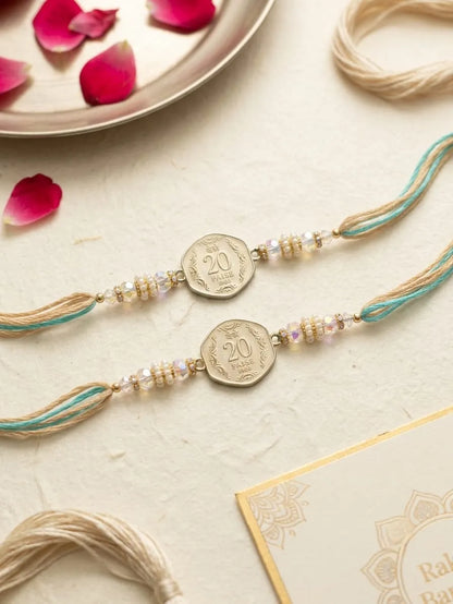 Two traditional Indian Rakhi strands with gold-colored coin charms, decorated with light blue and beige threads, arranged on a textured cream surface with pink rose petals and a gold card.