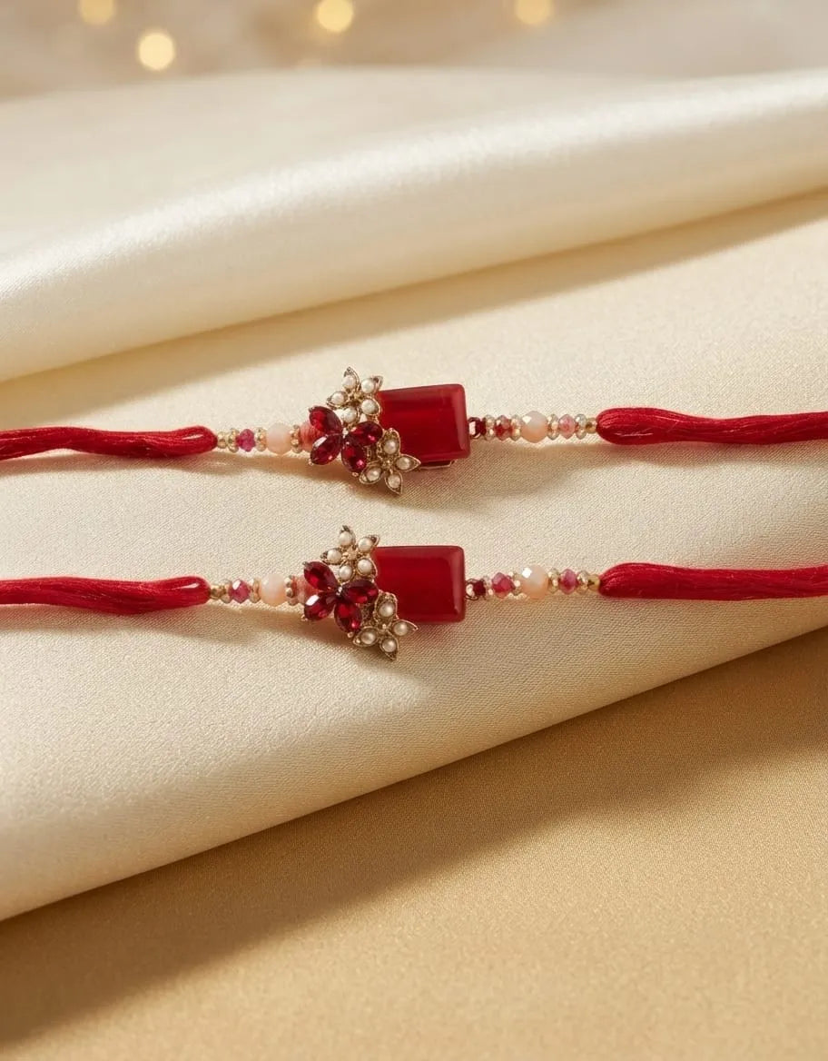 Two red thread Rakhi bracelets with red rectangular beads, floral embellishments, and small clear stones, displayed on draped cream satin fabric.