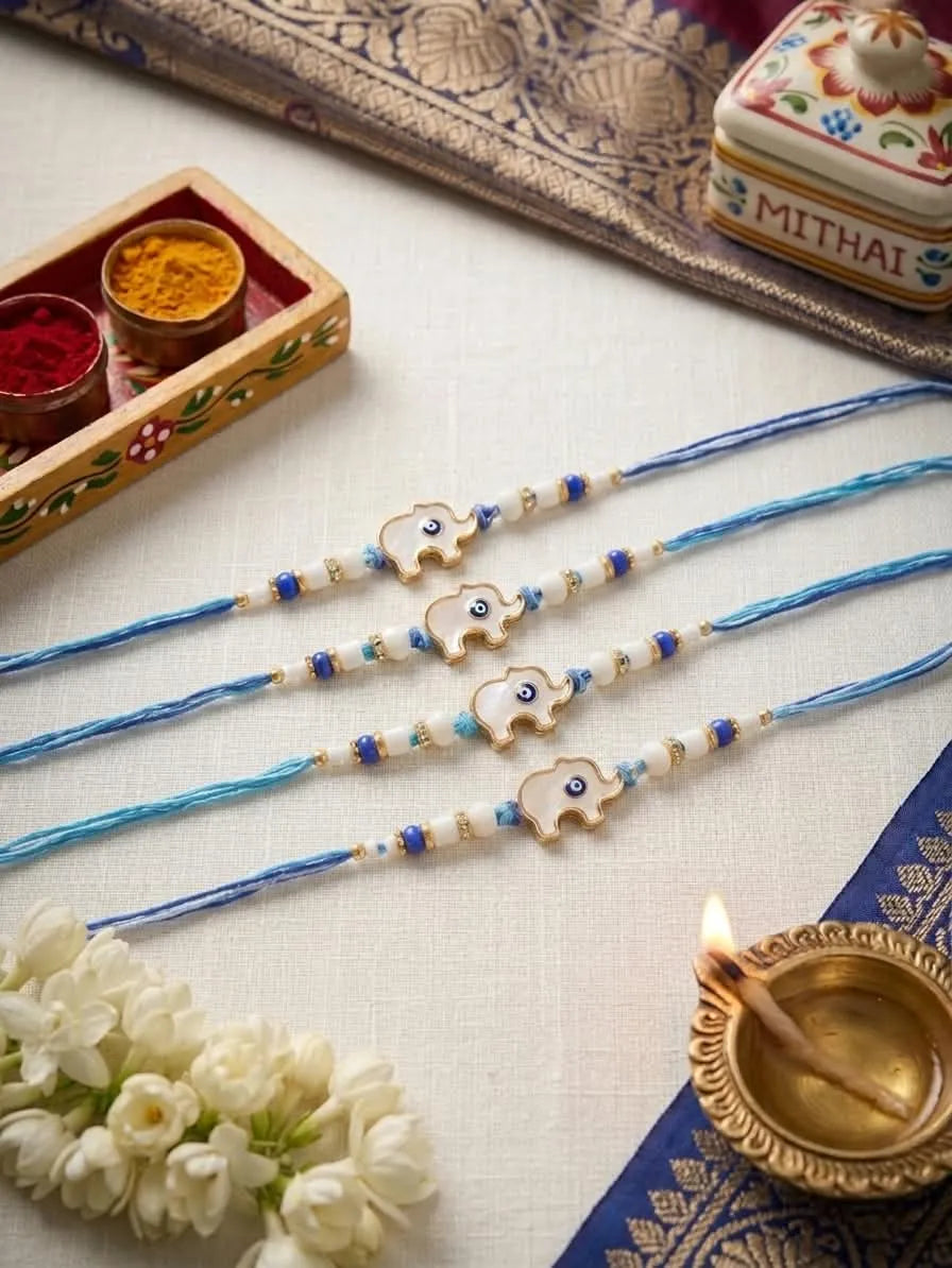 Four blue thread Rakhi bracelets with small elephant charms and white/blue beads, arranged on a white fabric surface with decorative props including flowers, a brass diya, a match box, and a Mithai box.