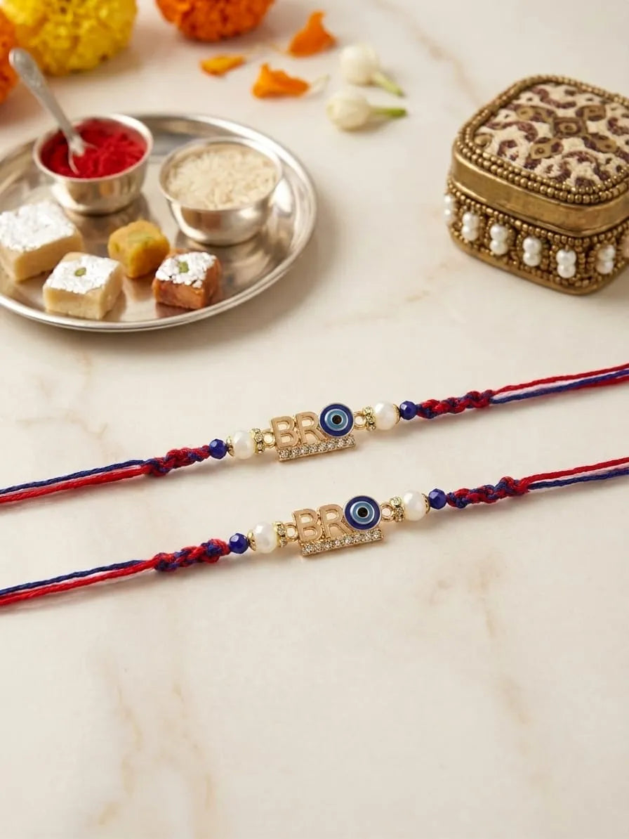Two traditional Indian Rakhi string bracelets with gold-toned evil eye charms and white pearl beads, displayed on a light marble surface with festive sweets and decorative items in the background.