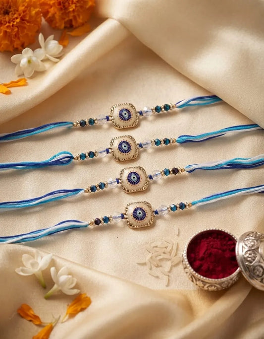 Four blue and white braided Rakhi strings with central silver-toned evil eye charms, arranged on draped cream satin fabric with decorative flowers and a red powder bowl.