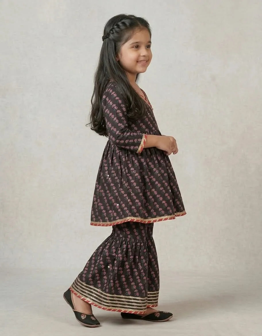 A young girl modeling a dark brown and pink printed kurta and flared pants set, standing against a neutral textured beige background.