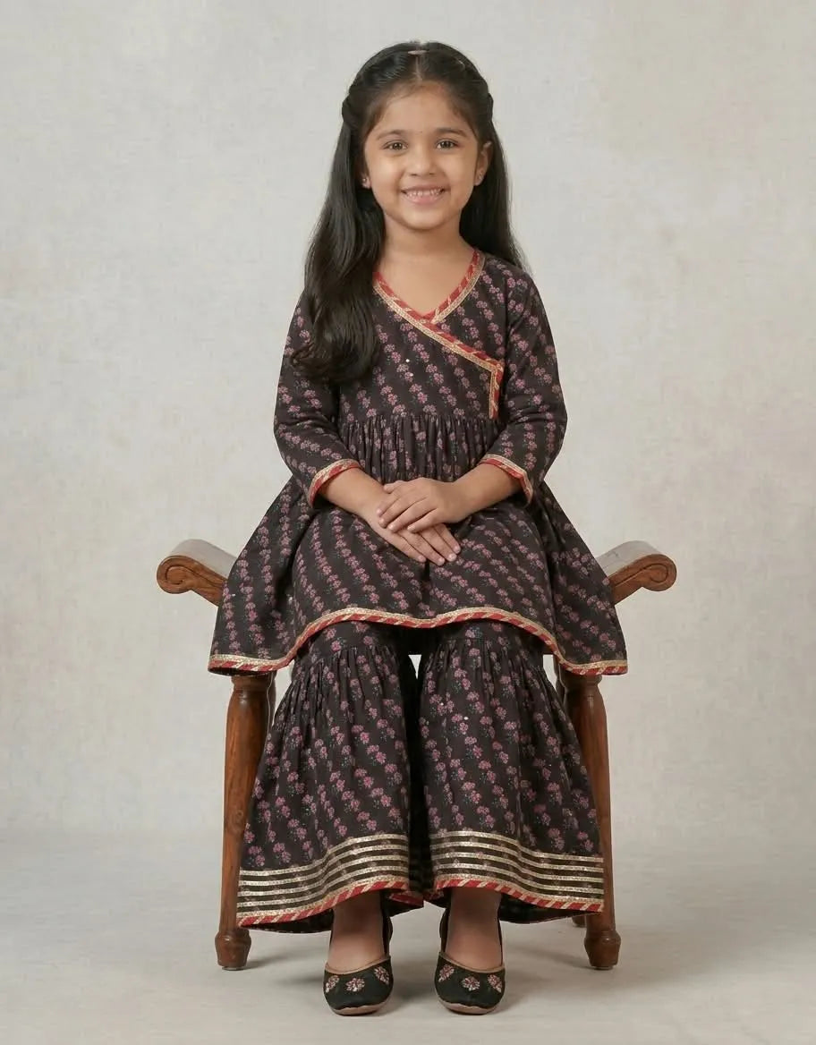 A young girl with long black hair sits on a wooden chair, wearing a dark brown printed salwar kameez set with pink floral motifs and red and gold borders. The set includes a long-sleeve tunic and matching wide-leg pants, photographed against a neutral beige studio backdrop.