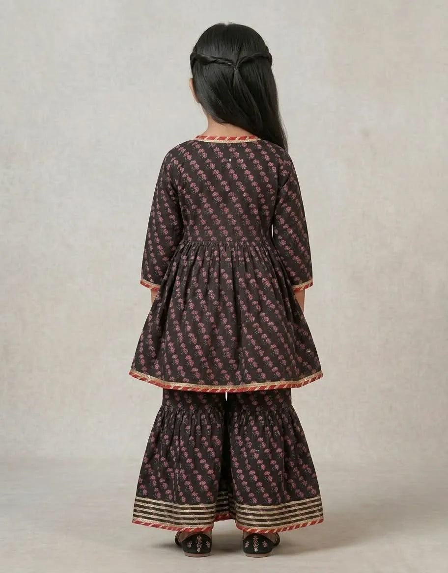 A child model shown from the back wearing a dark brown and pink patterned kurta and flared pants set with red and gold borders, set against a neutral studio backdrop.