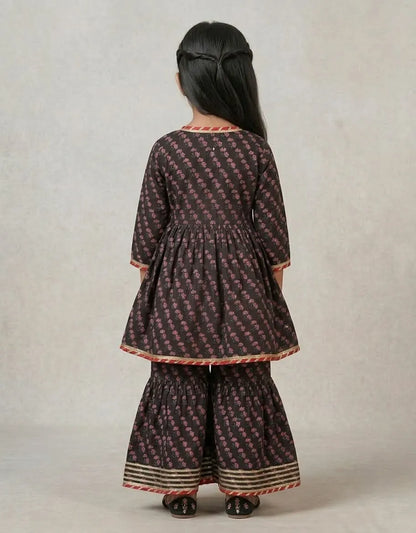 A child model shown from the back wearing a dark brown and pink patterned kurta and flared pants set with red and gold borders, set against a neutral studio backdrop.