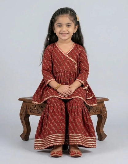 A young girl with long dark hair sits on a carved wooden bench, wearing a rust-red printed salwar kameez outfit with gold zari borders. The background is a solid light gray studio backdrop.