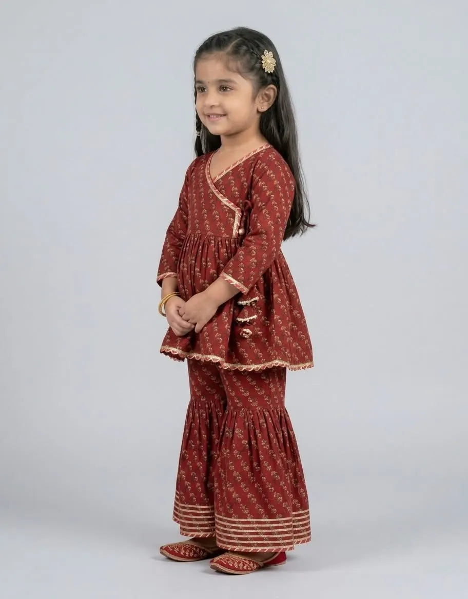 A young girl modeling a rust-red printed kurta and flared pants set with gold trim, standing against a light gray studio background.