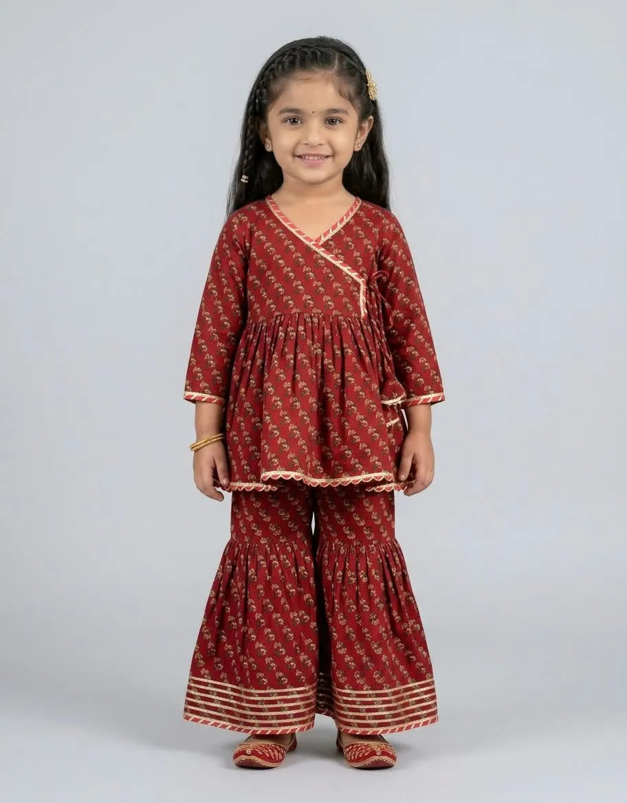 A young girl modeling a rust red printed kurta and flared pants set with beige borders, shot against a solid light gray background.
