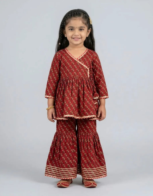 A young girl modeling a rust red printed kurta and flared pants set with beige borders, shot against a solid light gray background.