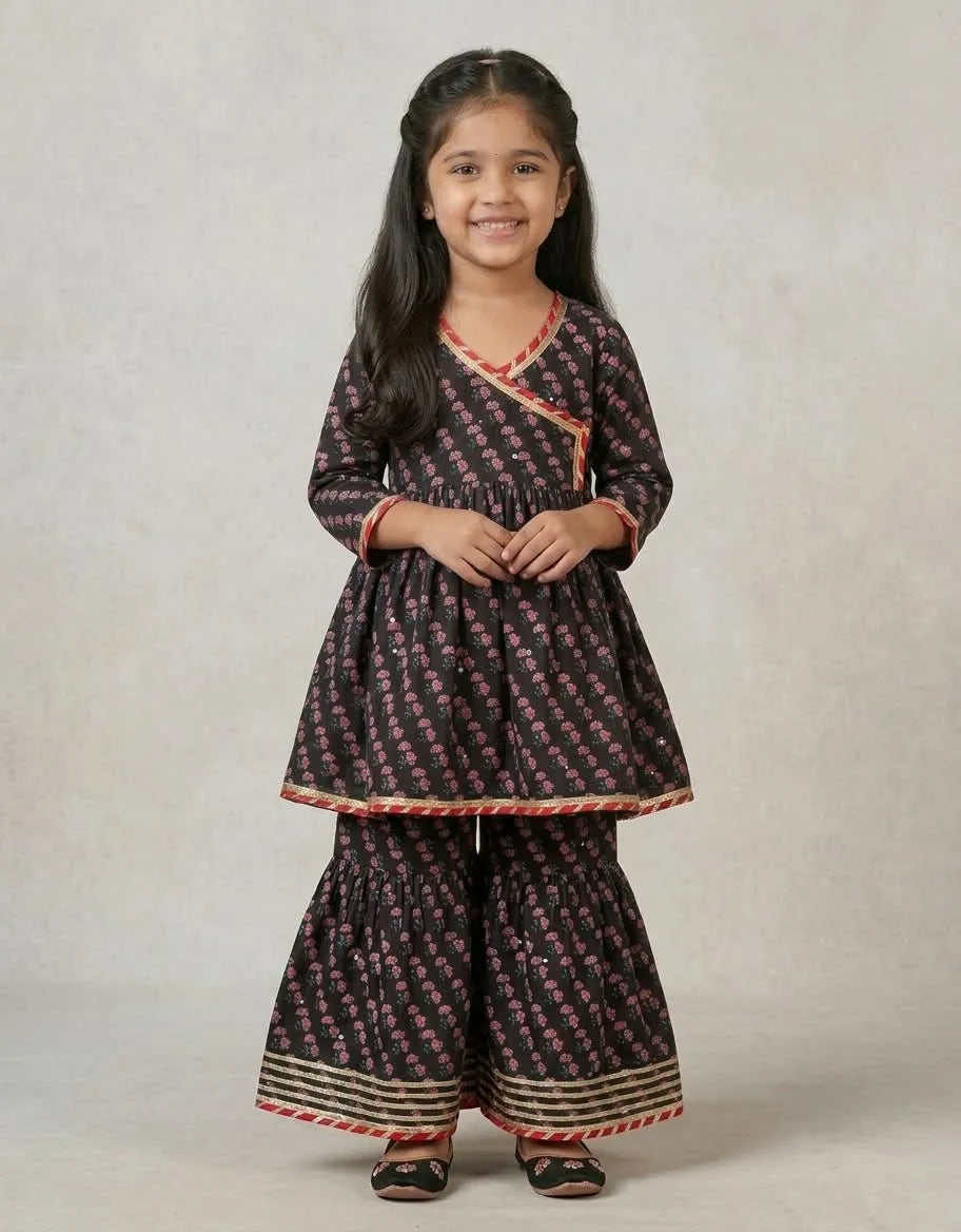 A young girl modeling a dark brown and pink floral print kurta and flared trouser set with red and gold border details, standing against a neutral light gray background.