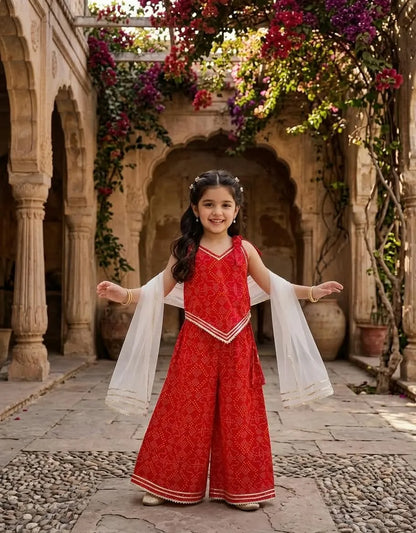 A young girl stands in a stone courtyard with arches and hanging flowers, wearing a red patterned sleeveless outfit with wide legs and white sheer draped sleeves.