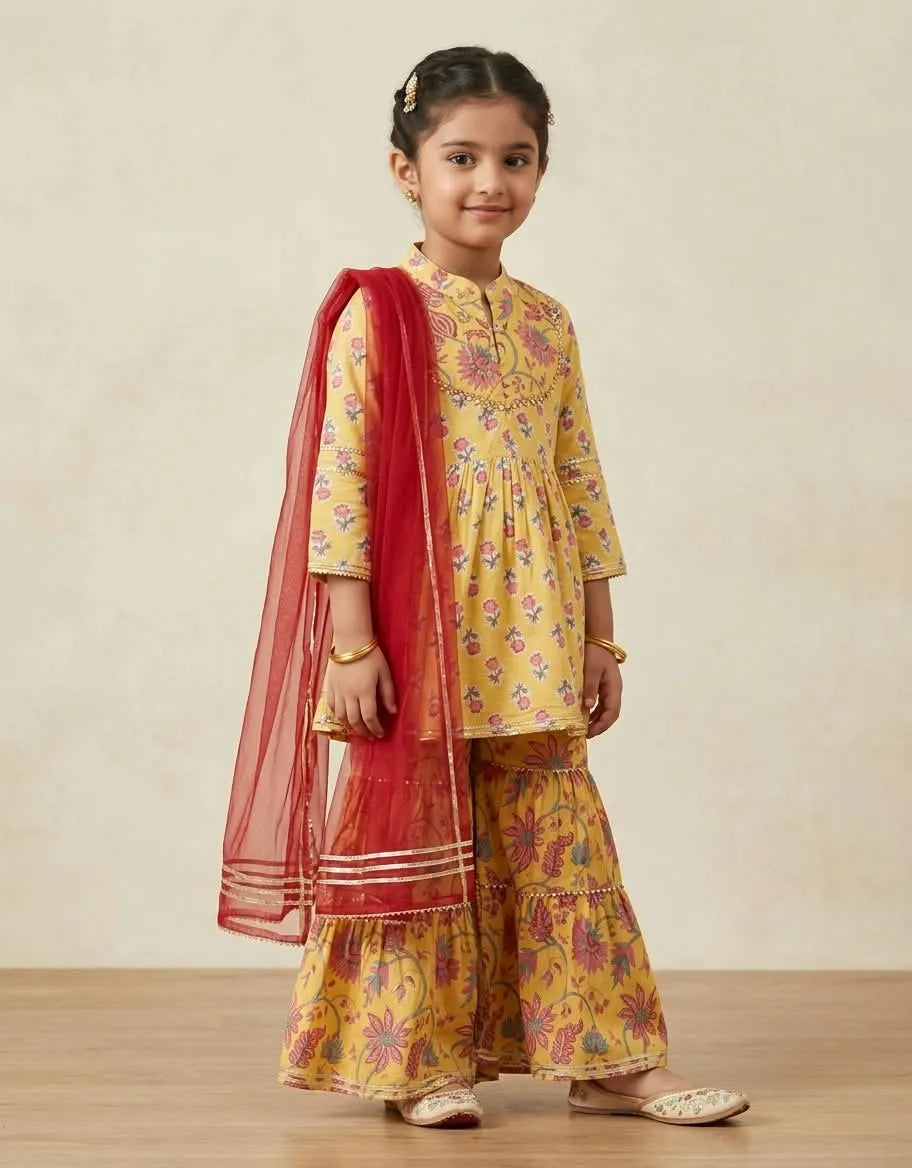 A young girl modeling a yellow floral printed kurta and flared pants set with a red dupatta, standing against a neutral beige background.