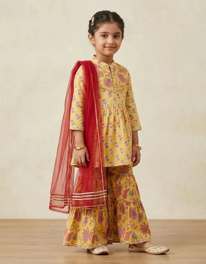 A young girl modeling a yellow floral printed kurta and flared pants set with a red dupatta, standing against a neutral beige background.