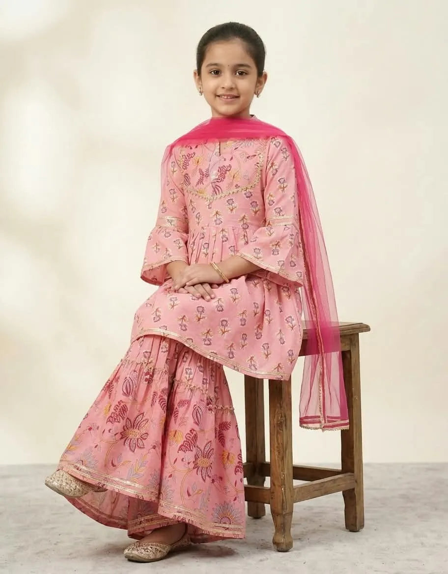 A young girl wearing a pink printed traditional outfit with a matching sheer dupatta, sitting on a wooden stool against a plain beige studio background.